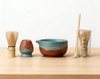 May include: A matcha tea set featuring a bamboo whisk, a ceramic bowl with a blue and brown glaze, a small ceramic holder, a bamboo scoop, and a container with a bamboo whisk. The set is arranged on a wooden surface.