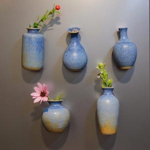 May include: Five blue ceramic vases of varying shapes, each with a speckled finish. Some vases hold flowers and greenery. The vases have a gradient color scheme, transitioning from blue to a yellowish-brown at the base.