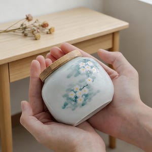 May include: A small, round, ceramic jar with a wooden lid. The jar is light blue with a floral design in white and green. The jar is held in two hands.