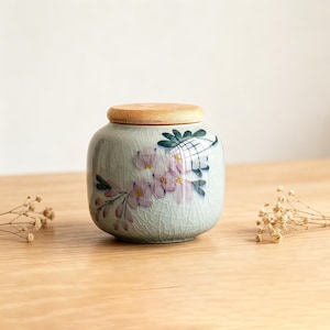 May include: A round ceramic jar with a light blue crackle glaze finish, featuring a painted floral design in pink and dark green. The container is topped with a circular, natural-coloured wooden lid that sits flush on the top opening.