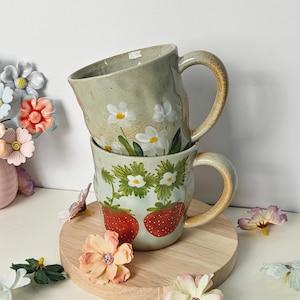 Könnte beinhalten: Zwei gestapelte Keramikbecher mit floralen Designs. Der obere Becher zeigt weiße Blumen mit gelben Zentren, während der untere Becher Erdbeeren und weiße Blumen zeigt. Beide Becher haben beige Griffe und stehen auf einer runden Holzbasis.