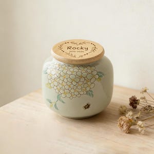 May include: A ceramic urn with a pale green body, decorated with a floral pattern and a wooden lid. The lid is engraved with the name "Rocky" and the years "2015-2026". The urn is designed for memorial purposes.