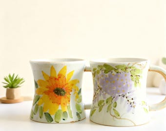 May include: Two ceramic mugs with floral patterns. One mug displays a yellow and orange sunflower, the other purple wisteria. Both mugs have a white base and curved handles, ideal for hot beverages.