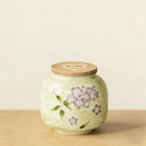 Puede incluir: Una urna de cerámica verde claro con tapa de madera. La urna está decorada con diseños florales morados y hojas verdes. La tapa está grabada con el nombre "Rocky" y los años "2015-2026".