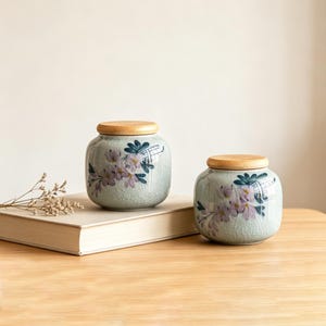 May include: Two ceramic jars with wooden lids. The jars are light green with a floral design in purple and blue. The jars are sitting on a book on a wooden table.