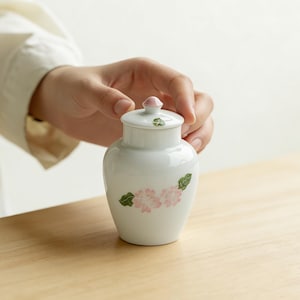 Puede incluir: Un jarrón de cerámica blanco con tapa, con un diseño de flores rosas y hojas verdes. El jarrón tiene un pequeño pomo rosa en la tapa. El jarrón es sostenido por una persona.