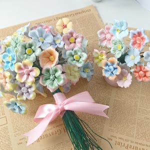 Puede incluir: Un ramo de flores de cerámica en colores pastel, azules, rosas, amarillas y verdes, atado con una cinta rosa. También se ve un arreglo más pequeño de flores similares.