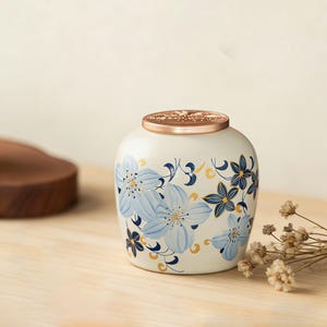 Puede incluir: Una urna de cerámica blanca con tapa de bronce y diseño floral. La urna presenta flores azul claro con detalles en azul oscuro y dorado. La urna está sobre una superficie de madera clara.