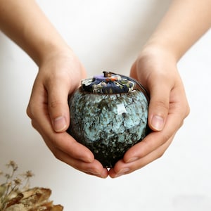 Puede incluir: Un pequeño jarrón de cerámica redondo con un esmalte moteado verde azulado y negro. El jarrón se sostiene con dos manos y tiene una tapa estampada con un acento púrpura. El jarrón parece ser un artículo decorativo o un pequeño recipiente de almacenamiento.