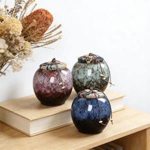 Puede incluir: Tres tarros de cerámica con tapas, cada uno de un color diferente: burdeos, verde azulado y azul marino. Los tarros tienen un acabado brillante y están adornados con tapas de tela estampada y lazos de cuerda. Están sobre una superficie de madera.