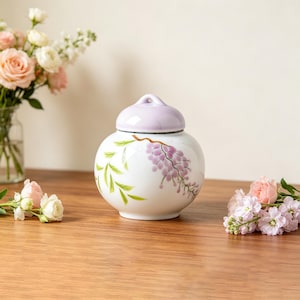 May include: A white ceramic jar with a rounded body and a matching light purple lid. The jar features a hand-painted design of purple wisteria flowers and green leaves on its exterior. The surface is smooth and glossy, with a small handle on the lid.