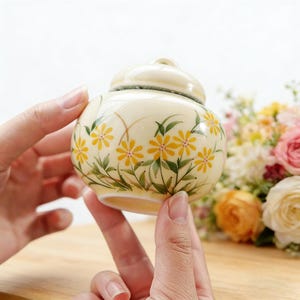 Könnte beinhalten: Ein kleines, rundes Keramikgefäß mit Deckel, verziert mit gelben, gänseblümchenartigen Blumen und grünen Blättern. Das Gefäß hat eine cremefarbene Basis und einen passenden Deckel. Das Blumendesign ist handgemalt.