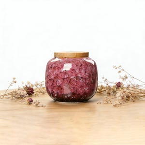 Puede incluir: Un tarro redondo de cerámica con un exterior rojo oscuro texturizado y una tapa de madera marrón claro. El tarro se encuentra sobre una superficie de madera marrón claro, con flores secas en el fondo. El tarro tiene un acabado brillante.