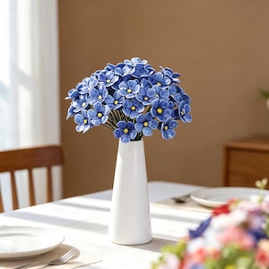 Könnte beinhalten: Ein Strauß blauer Keramikblumen mit gelben Zentren in einer weißen Vase. Die Vase steht auf einer weißen Tischdecke, mit einem Teller und Besteck. Die Blumen sind der Blickfang.