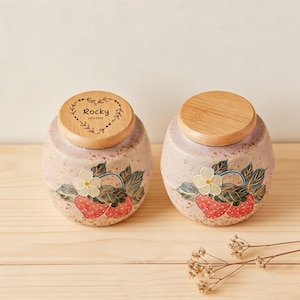 May include: Two ceramic jars with wooden lids. The jars are a speckled pink with a strawberry design. One lid has the name "Rocky" and the years "2015-2026" engraved. The jars are on a light wood surface.