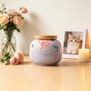 以下が含まれることがあります： 淡い紫色のまだら模様の仕上げが施された丸い陶器製の瓶。浮き彫りになったピンクの牡丹の花と小さな青い蝶のデザインが特徴です。瓶の上部には、開口部にぴったりと収まるナチュラルカラーの円形木製蓋が付いています。