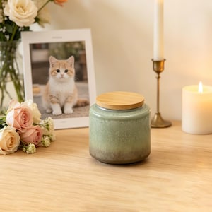 May include: A cylindrical ceramic candle jar with a light green to dark brown gradient glaze and a smooth, circular wooden lid. The jar features a speckled texture throughout the surface, providing a distinct contrast between the lighter top and darker base.