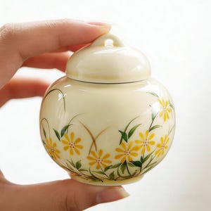 Puede incluir: Un pequeño tarro de cerámica redondo de color crema con tapa a juego. El tarro está decorado con flores amarillas en forma de margarita y hojas verdes. El diseño floral está pintado a mano, añadiendo un toque artístico. El tarro es sostenido por una mano.
