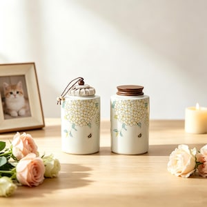 May include: Two cylindrical ceramic canisters featuring a white base with a yellow and green hydrangea floral pattern and small bee illustrations. One canister has a fabric-covered lid with a brown cord, while the other has a dark wooden lid.