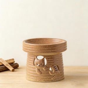 May include: A tan ceramic cat-themed oil warmer. The warmer has a cylindrical shape with a wide top and a cat design on the side. The cat is partially visible through a circular opening. The warmer is on a wooden surface.