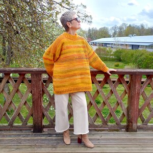 Puede incluir: Una persona con un jersey de rayas amarillas y naranjas con cuello redondo y mangas largas. También lleva pantalones anchos color crema y botines color canela. El fondo muestra una terraza de madera, árboles y un edificio a lo lejos.