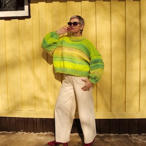 May include: A cropped, hand-knitted sweater in shades of green and yellow, paired with cream-colored wide-leg pants and red shoes. The sweater features a round neck and long sleeves. The background is a yellow wall.