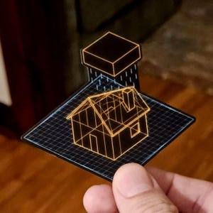 Puede incluir: Una pegatina transparente con un diseño de casa geométrica con contorno naranja. La casa está sobre una cuadrícula negra, con un cuadrado encima. La pegatina está sujeta por una mano.