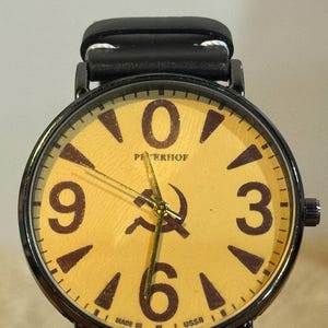 Peut inclure: Une montre-bracelet avec un boîtier en métal noir et un bracelet en cuir noir. Le cadran est jaune clair avec de grands chiffres marron foncé à 12, 3, 6 et 9. Il présente un symbole marteau et faucille, le texte PETERHOF et MADE IN USSR.