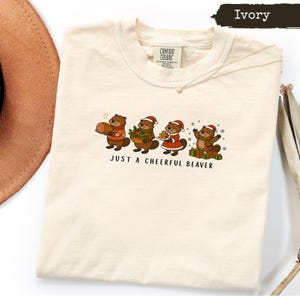 May include: An ivory t-shirt featuring a graphic of four cartoon beavers wearing Christmas hats and festive clothing. The beavers are holding holiday items including a ham, a bowl of greens, a cookie, and gifts. The text reads, JUST A CHEERFUL BEAVER.