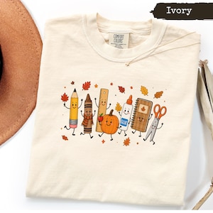 May include: An ivory t-shirt featuring a graphic print of anthropomorphic school supplies including a pencil, crayon, ruler, pumpkin, glue bottle, notebook, and scissors, surrounded by small autumn leaves. The shirt label reads Comfort Colors.