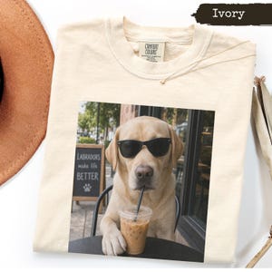 May include: An ivory t-shirt featuring a graphic print of a yellow Labrador retriever wearing black sunglasses and drinking an iced coffee from a clear plastic cup. The print includes a chalkboard sign that reads Labradors make life BETTER.