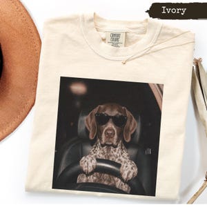 Op de afbeelding: Een ivoorkleurig T-shirt met ronde hals en een grafische print van een bruin met wit gevlekte hond die een zwarte zonnebril draagt terwijl hij op de bestuurdersstoel van een auto zit. Het T-shirt heeft een label van het merk Comfort Colors.