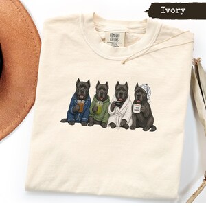 Op de afbeelding: Een ivoorkleurig t-shirt met een afbeelding van vier zwarte Cane Corso honden. De honden dragen pyjama's, een hoodie, een badjas en een slaapmuts, terwijl ze koffie- en theedrankjes vasthouden. Op één mok staat de tekst I NEED COFFEE.