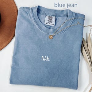 May include: A blue jean coloured Comfort Colors t-shirt with the word "NAH" printed in white. A gold necklace with a pendant is draped across the shirt. A brown hat and dried plant are in the background.