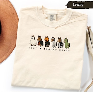 May include: An ivory t-shirt featuring a graphic of six horses in various Halloween costumes, including a ghost, vampire, pumpkin, mummy, skeleton, and zombie. The text below the horses reads, "JUST A SPOOKY HORSE." The shirt has a Comfort Colors label.
