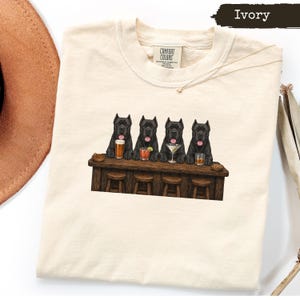 Könnte beinhalten: Ein elfenbeinfarbenes T-Shirt mit einer Grafik von vier schwarzen Cane Corso Hunden, die an einer Holzbar sitzen und jeweils ein anderes Getränk vor sich haben. Das Shirt hat einen Rundhalsausschnitt und ein Comfort Colors Etikett. Text Ivory sichtbar.