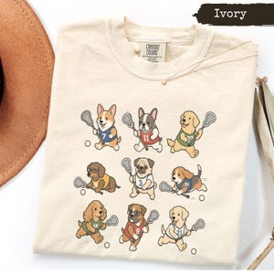 Peut inclure: Un t-shirt ivoire avec une impression graphique de neuf chiens de dessin animé jouant à la crosse. Chaque chien porte un maillot numéroté de différentes couleurs et tient une crosse. Le t-shirt porte l'étiquette Comfort Colors au col.