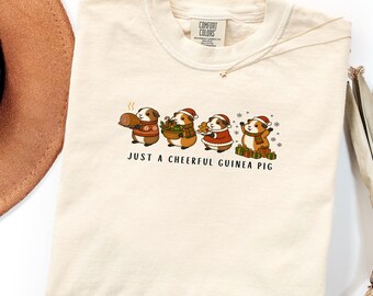 May include: An ivory t-shirt featuring a graphic of four guinea pigs wearing Santa hats and holiday attire. The guinea pigs are holding a ham, a bowl of greens, a cookie, and gifts. The text below reads, JUST A CHEERFUL GUINEA PIG.