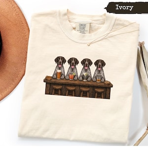 May include: An ivory-colored t-shirt featuring a graphic of four German Shorthaired Pointer dogs sitting at a wooden bar. Each dog has a different alcoholic beverage in front of it, including a beer, a cocktail, a martini, and a whiskey glass.