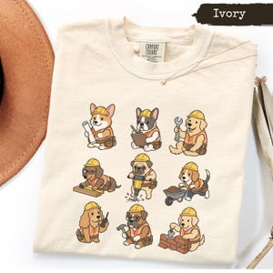 May include: An ivory t-shirt featuring a graphic print of nine cartoon dogs dressed as construction workers in yellow hard hats and orange safety vests. The dogs are depicted performing various tasks with tools like a wrench, hammer, and jackhammer.