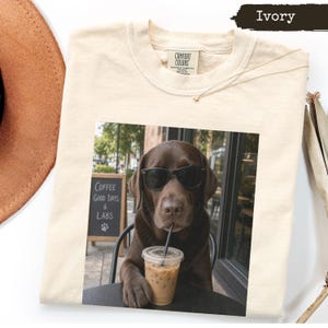 May include: An ivory crew neck t-shirt featuring a graphic print of a chocolate Labrador dog wearing black sunglasses and holding a plastic cup with a straw. The dog is sitting at a table next to a chalkboard sign that reads Coffee Good Days & Labs.
