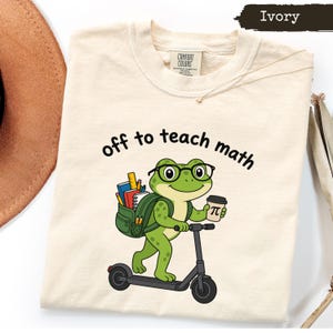 May include: An ivory t-shirt featuring a graphic of a green frog wearing black glasses, carrying a backpack filled with books and pencils, riding a black scooter, and holding a coffee cup with a pi symbol. The text reads off to teach math.