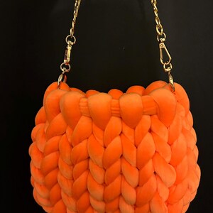 Puede incluir: Bolso tejido a mano de color naranja brillante con una correa de cadena dorada. El bolso está hecho de un material suave y afelpado y presenta un diseño trenzado. La cadena está sujeta con cierres dorados. El bolso está sobre un fondo negro.