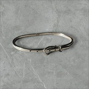 May include: A silver-toned metal bangle bracelet designed to resemble a miniature belt. The bracelet features a thin, smooth band with a functional-style buckle closure, a metal loop, and small holes along the strap to mimic a belt's appearance.