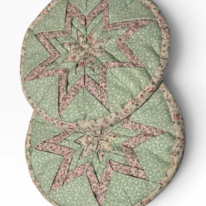 May include: Two circular quilted pot holders featuring a central star pattern. The fabric displays a light green background with a small white floral print, accented by a pink and green floral pattern on the star points and the outer edge binding.