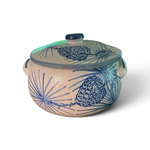 May include: A round, off-white ceramic lidded pot featuring a hand-painted blue design of pinecones and pine needles. The vessel includes two small side handles and a matching lid with a central knob, showcasing a rustic, speckled texture.