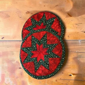 May include: Two round quilted potholders featuring a central eight-pointed star design. The fabric displays a pattern of red and dark green sections with small white floral accents. The edges are finished with a dark green binding trim.