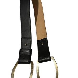 May include: A black belt featuring a smooth exterior and a tan interior lining. The belt is secured with two large, asymmetrical gold-tone metal loops at the ends. One of the gold loops is engraved with the text Oscar de la Renta in cursive script.