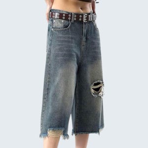 Op de afbeelding: Blauwe denim culottes met distressed details, met een gescheurde knie en gerafelde zoom. De broek wordt gedragen met een bruine riem versierd met zilverkleurige stervormige studs.