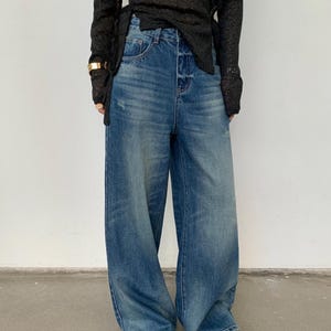 May include: Blue denim wide-leg jeans with a faded wash. The jeans have a high waist and a relaxed fit. The denim pants have a classic five-pocket design and a button and zipper closure. The jeans are worn with a black top.